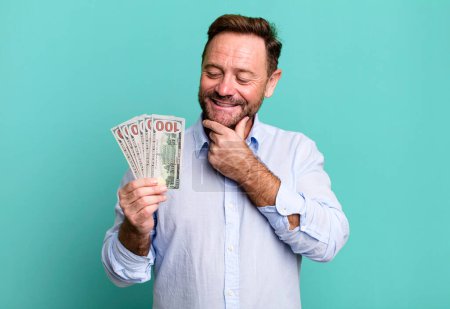 Foto de Middle age man smiling with a happy, confident expression with hand on chin. dollar banknotes concept - Imagen libre de derechos