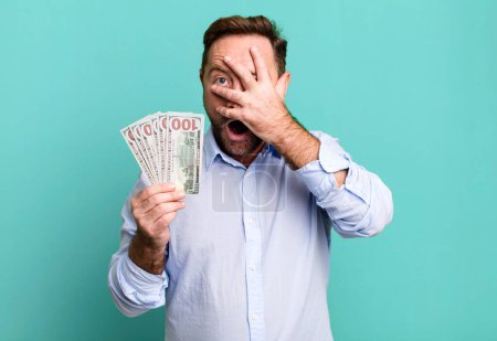 Foto de Middle age man looking shocked, scared or terrified, covering face with hand. dollar banknotes concept - Imagen libre de derechos
