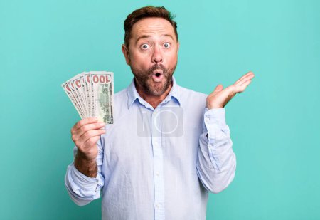 Foto de Middle age man looking surprised and shocked, with jaw dropped holding an object. dollar banknotes concept - Imagen libre de derechos