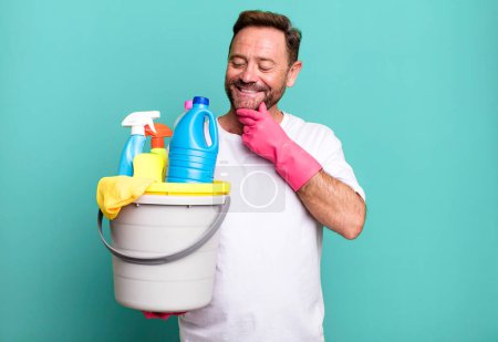 Photo for Middle age man smiling with a happy, confident expression with hand on chin. housekeeper with clean products - Royalty Free Image