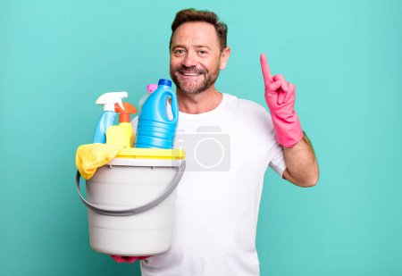 Foto de Hombre de mediana edad sonriendo y buscando amigable, mostrando el número uno. ama de llaves con productos limpios - Imagen libre de derechos