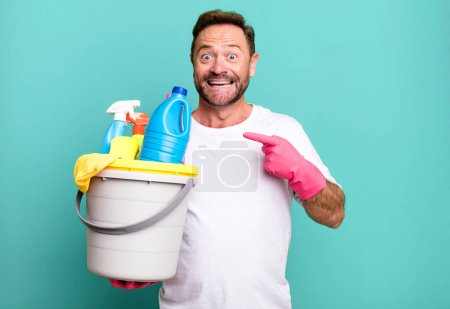 Photo for Middle age man looking excited and surprised pointing to the side. housekeeper with clean products - Royalty Free Image