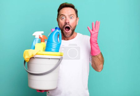 Photo for Middle age man feeling extremely shocked and surprised. housekeeper with clean products - Royalty Free Image