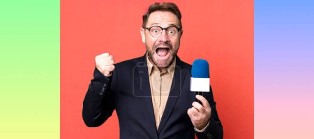 Foto de Hombre de mediana edad sintiéndose sorprendido, riendo y celebrando el éxito. periodista y un concepto de micrófono - Imagen libre de derechos