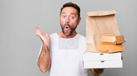 Téléchargez les photos : Homme d'âge moyen regardant surpris et choqué, avec la mâchoire tombée tenant un objet. livraison et fast food concept à emporter - en image libre de droit