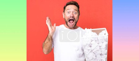 Photo for Middle age man feeling happy and astonished at something unbelievable. paper balls trash basket - Royalty Free Image