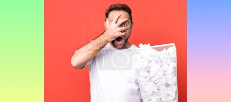 Foto de Hombre de mediana edad que parece sorprendido, asustado o aterrorizado, cubriendo la cara con la mano. bolas de papel papelera - Imagen libre de derechos