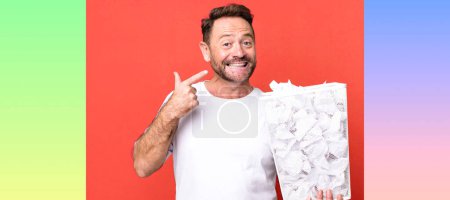 Photo for Middle age man smiling confidently pointing to own broad smile. paper balls trash basket - Royalty Free Image