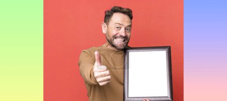 Photo for Middle age man feeling proud,smiling positively with thumbs up with an empty frame - Royalty Free Image
