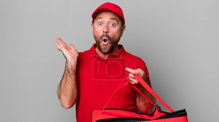 Téléchargez les photos : Homme d'âge moyen regardant surpris et choqué, avec la mâchoire tombée tenant un objet. livreur de pizza - en image libre de droit