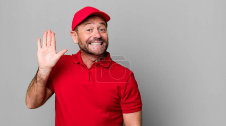 Téléchargez les photos : Homme d'âge moyen souriant joyeusement, main agitant, vous accueillant et vous saluant. employé de l'entreprise - en image libre de droit