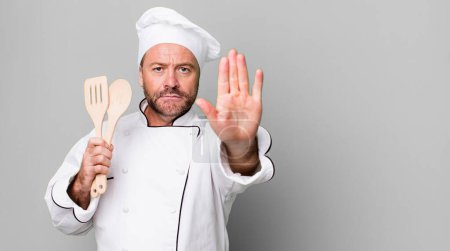 Foto de Hombre de mediana edad que se ve serio mostrando la palma abierta haciendo gesto de parada. concepto de chef y herramientas - Imagen libre de derechos