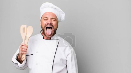 Foto de Hombre de mediana edad con actitud alegre y rebelde, bromeando y sacando la lengua. concepto de chef y herramientas - Imagen libre de derechos