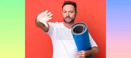 Photo for Middle age man feeling cross,showing thumbs down. with a yoga matt. fitness concept - Royalty Free Image