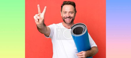 Téléchargez les photos : Homme d'âge moyen souriant et regardant heureux, geste de victoire ou de paix. avec un tapis de yoga. concept de fitness - en image libre de droit