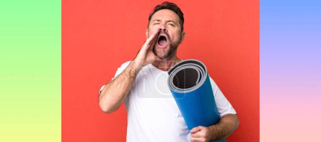 Photo for Middle age man feeling happy,giving a big shout out with hands next to mouth. with a yoga matt. fitness concept - Royalty Free Image