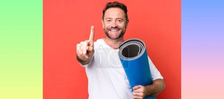 Foto de Hombre de mediana edad sonriendo orgullosamente y con confianza haciendo número uno. con un mate de yoga. concepto de fitness - Imagen libre de derechos