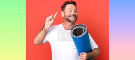 Téléchargez les photos : Homme d'âge moyen souriant et l'air amical, montrant numéro un. avec un tapis de yoga. concept de fitness - en image libre de droit
