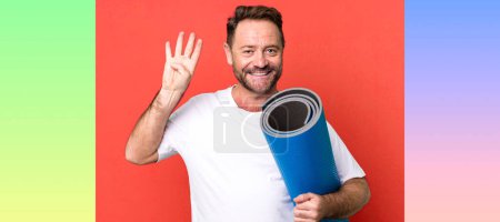 Téléchargez les photos : Homme d'âge moyen souriant et l'air amical, montrant le numéro quatre. avec un tapis de yoga. concept de fitness - en image libre de droit