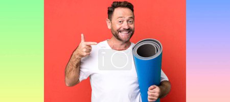 Foto de Hombre de mediana edad sonriendo con confianza apuntando a su propia sonrisa amplia. con un mate de yoga. concepto de fitness - Imagen libre de derechos