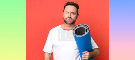 Téléchargez les photos : Homme d'âge moyen se sentant triste et pleurnichard avec un regard malheureux et pleurer. avec un tapis de yoga. concept de fitness - en image libre de droit