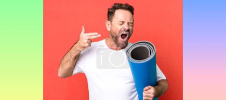 Foto de Hombre de mediana edad que parece infeliz y estresado, gesto suicida haciendo señal de arma. con un mate de yoga. concepto de fitness - Imagen libre de derechos
