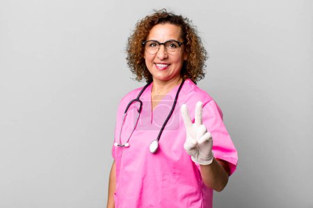 Foto de Mujer bastante de mediana edad sonriendo y buscando amigable, mostrando el número dos. concepto de enfermera - Imagen libre de derechos