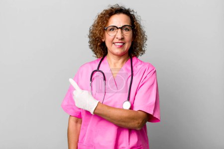 Photo for Pretty middle age woman smiling cheerfully, feeling happy and pointing to the side. nurse concept - Royalty Free Image