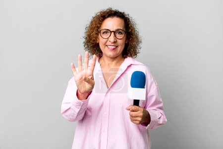 Foto de Bastante mujer de mediana edad sonriendo y buscando amigable, mostrando el número cuatro. presentador de televisión con un concepto de micrófono - Imagen libre de derechos