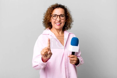 Foto de Bastante mujer de mediana edad sonriendo y buscando amigable, mostrando el número uno. presentador de televisión con un concepto de micrófono - Imagen libre de derechos