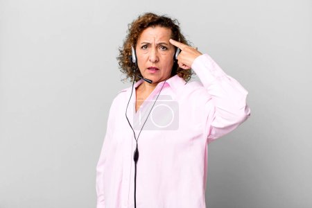 Photo for Pretty middle age woman feeling confused and puzzled, showing you are insane with a headset. telemarketing concept - Royalty Free Image