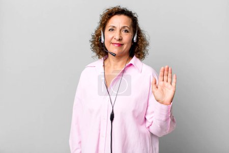 Foto de Bonita mujer de mediana edad sonriendo felizmente, saludando con la mano, dando la bienvenida y saludándote con un auricular. concepto de telemarketing - Imagen libre de derechos