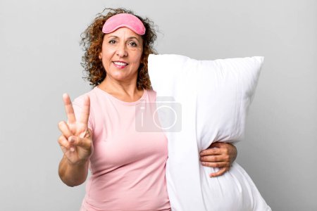 Foto de Bonita mujer de mediana edad sonriendo y luciendo feliz, haciendo gestos de victoria o paz usando pijamas ropa de noche y una almohada - Imagen libre de derechos
