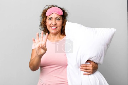 Foto de Mujer de mediana edad bonita sonriendo y buscando amigable, mostrando el número cuatro usando pijamas ropa de noche y una almohada - Imagen libre de derechos