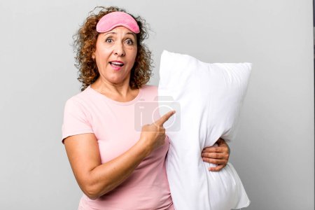 Foto de Mujer de mediana edad bonita mirando emocionado y sorprendido señalando al lado con pijama ropa de noche y una almohada - Imagen libre de derechos
