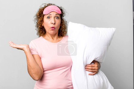 Foto de Mujer de mediana edad que parece sorprendida y conmocionada, con la mandíbula caída sosteniendo un objeto con pijama ropa de noche y una almohada - Imagen libre de derechos