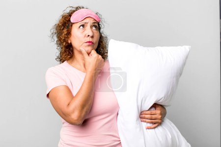Foto de Mujer de mediana edad pensando, sintiéndose dudosa y confundida usando pijamas ropa de noche y una almohada - Imagen libre de derechos