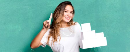 Photo for Hispanic pretty woman smiling and looking friendly, showing number one with white boxes packages - Royalty Free Image