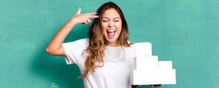 Photo for Hispanic pretty woman looking unhappy and stressed, suicide gesture making gun sign with white boxes packages - Royalty Free Image