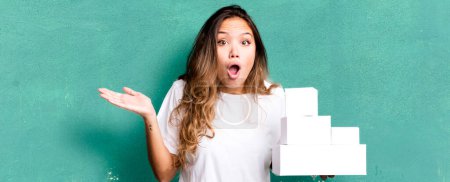 Foto de Hispanic pretty woman looking surprised and shocked, with jaw dropped holding an object with white boxes packages - Imagen libre de derechos