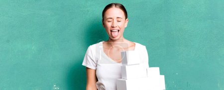 Foto de Caucasian pretty woman with cheerful and rebellious attitude, joking and sticking tongue out with white boxes packages - Imagen libre de derechos