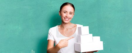 Téléchargez les photos : Caucasian pretty woman smiling cheerfully, feeling happy and pointing to the side with white boxes packages - en image libre de droit