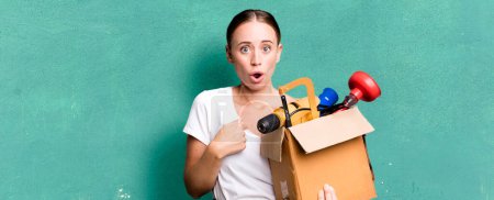 Photo for Caucasian pretty woman looking shocked and surprised with mouth wide open, pointing to self with a tool box. repair home concept - Royalty Free Image
