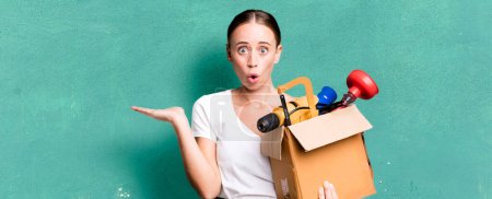 Foto de Caucasian pretty woman looking surprised and shocked, with jaw dropped holding an object with a tool box. repair home concept - Imagen libre de derechos