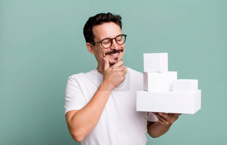 Foto de Adult man smiling with a happy, confident expression with hand on chin. blank boxes packaging concept - Imagen libre de derechos