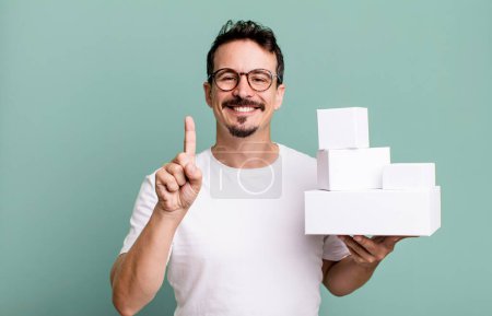 Téléchargez les photos : Adult man smiling and looking friendly, showing number one. blank boxes packaging concept - en image libre de droit