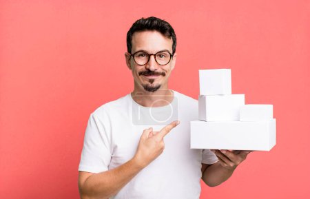 Téléchargez les photos : Adult man smiling cheerfully, feeling happy and pointing to the side. blank boxes packaging concept - en image libre de droit