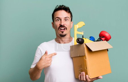 Foto de Adult man looking shocked and surprised with mouth wide open, pointing to self. with a toolbox. handyman concept - Imagen libre de derechos