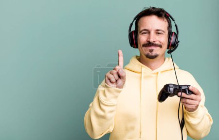 Foto de Adult man smiling and looking friendly, showing number one with headset and a control. gamer concept - Imagen libre de derechos
