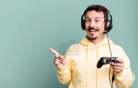 Photo for Adult man looking excited and surprised pointing to the side with headset and a control. gamer concept - Royalty Free Image